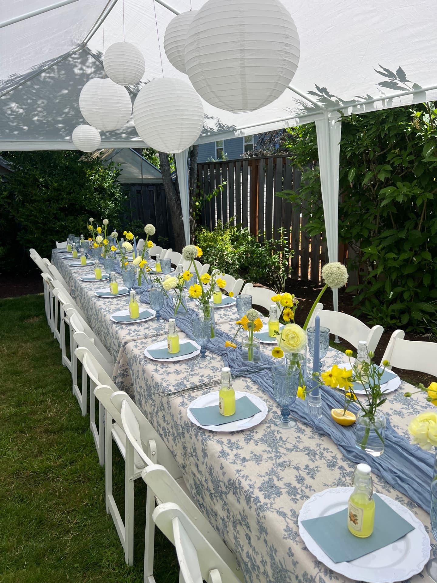 A long backyard table under a white tent set with blue toile linens, blue glassware, white plates, and yellow florals
