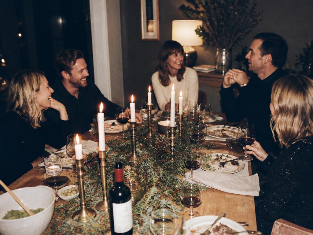 Friends gathered around a candlelit holiday dinner table with loose evergreen cuttings down the middle
