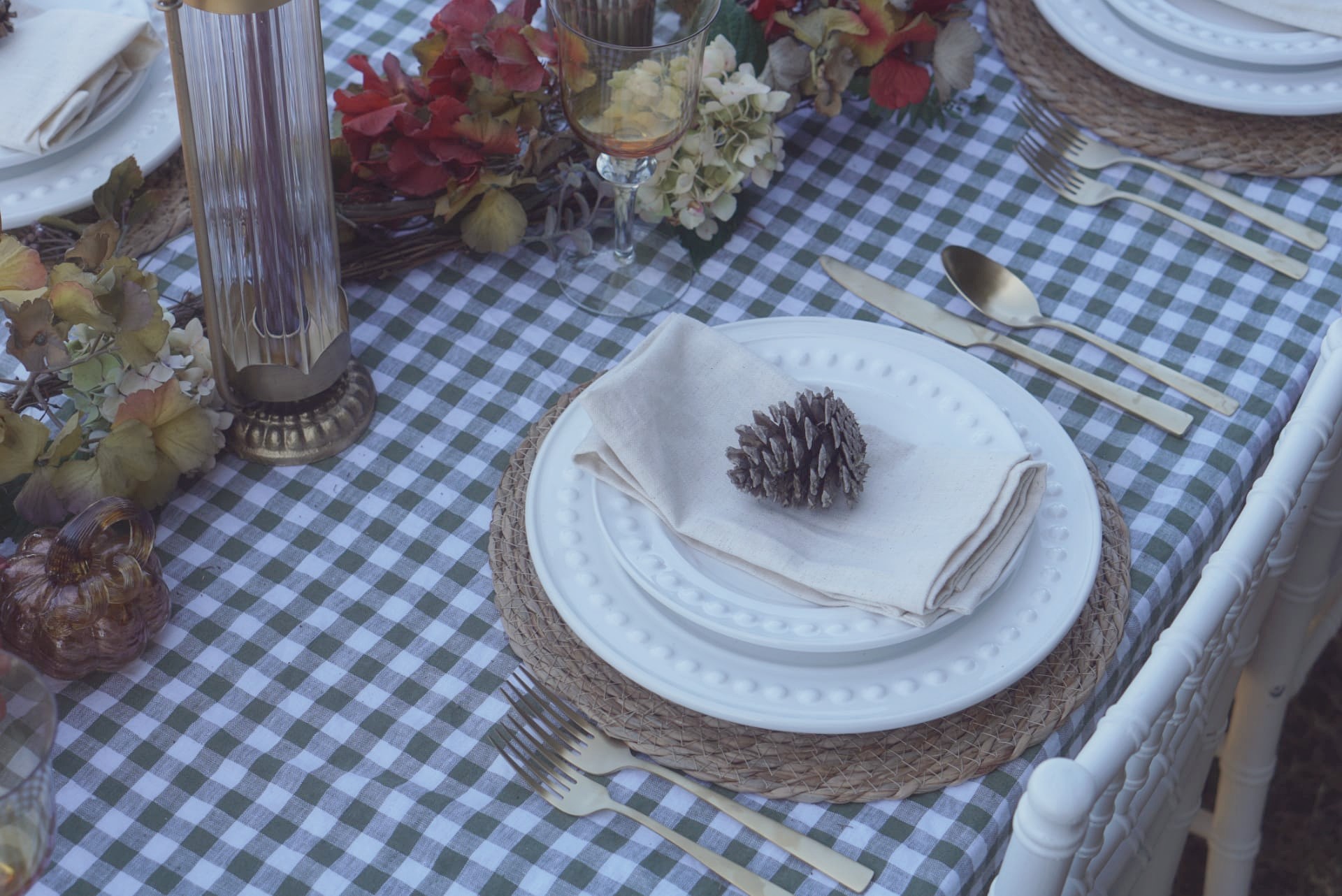Close-up place setting with woven charger, beaded white plates, linen napkin, pinecone, and gold flatware
