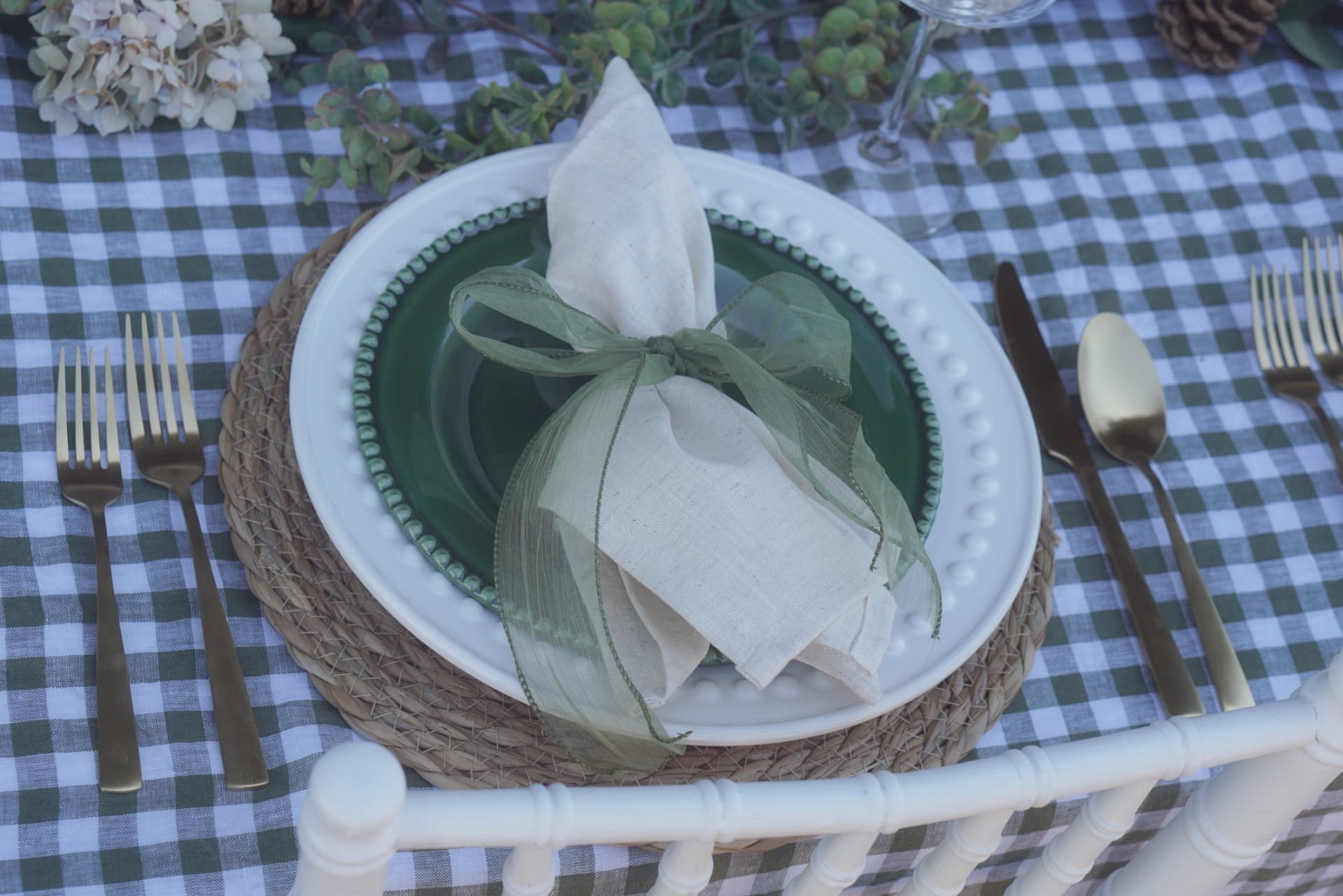 Close-up place setting with green gingham linens, green beaded plate, woven charger, linen napkin tied with sheer green ribbon, and gold flatware