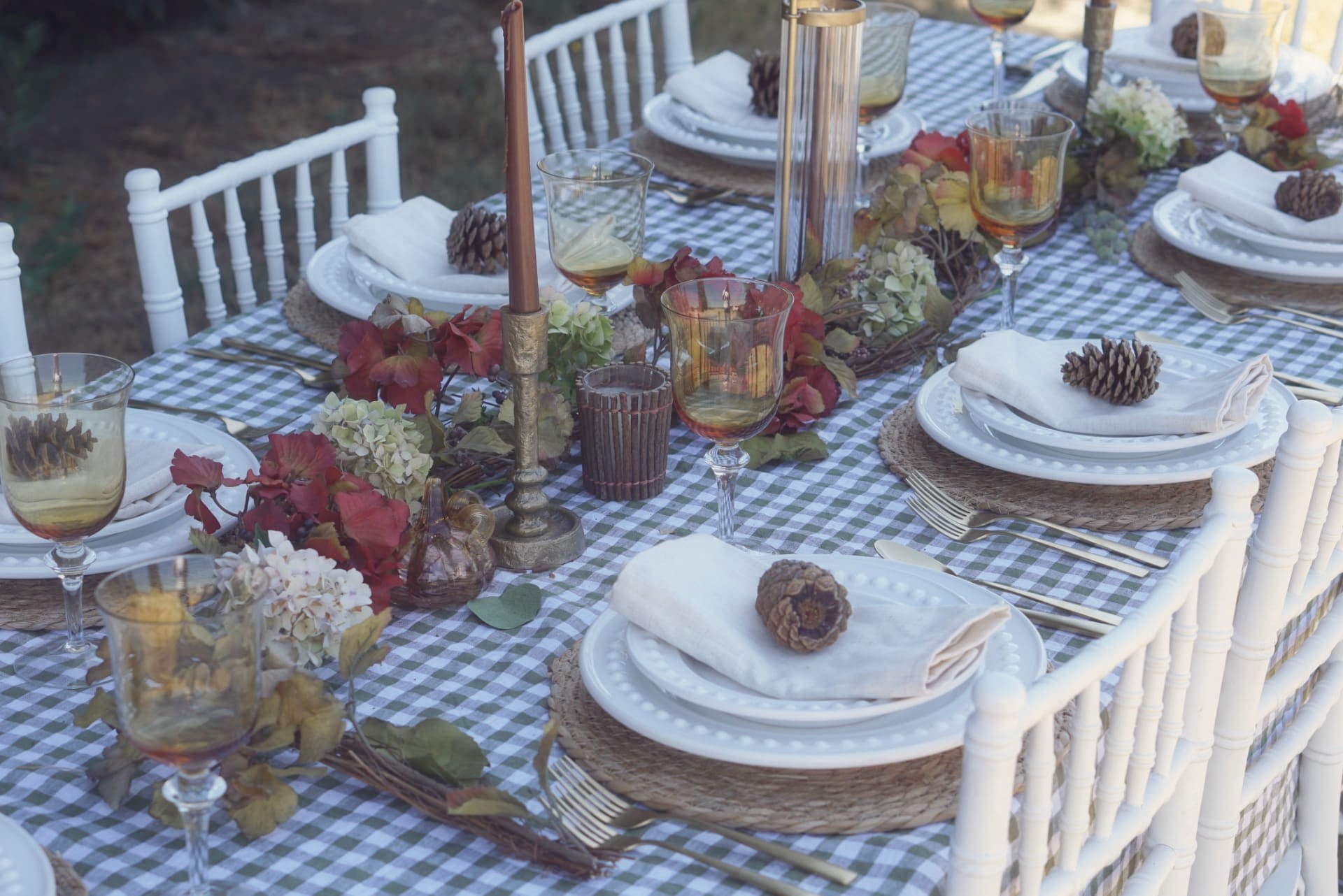A fall harvest tablescape with green gingham linens, beaded white plates, woven chargers, amber glassware, pinecones, and autumn foliage